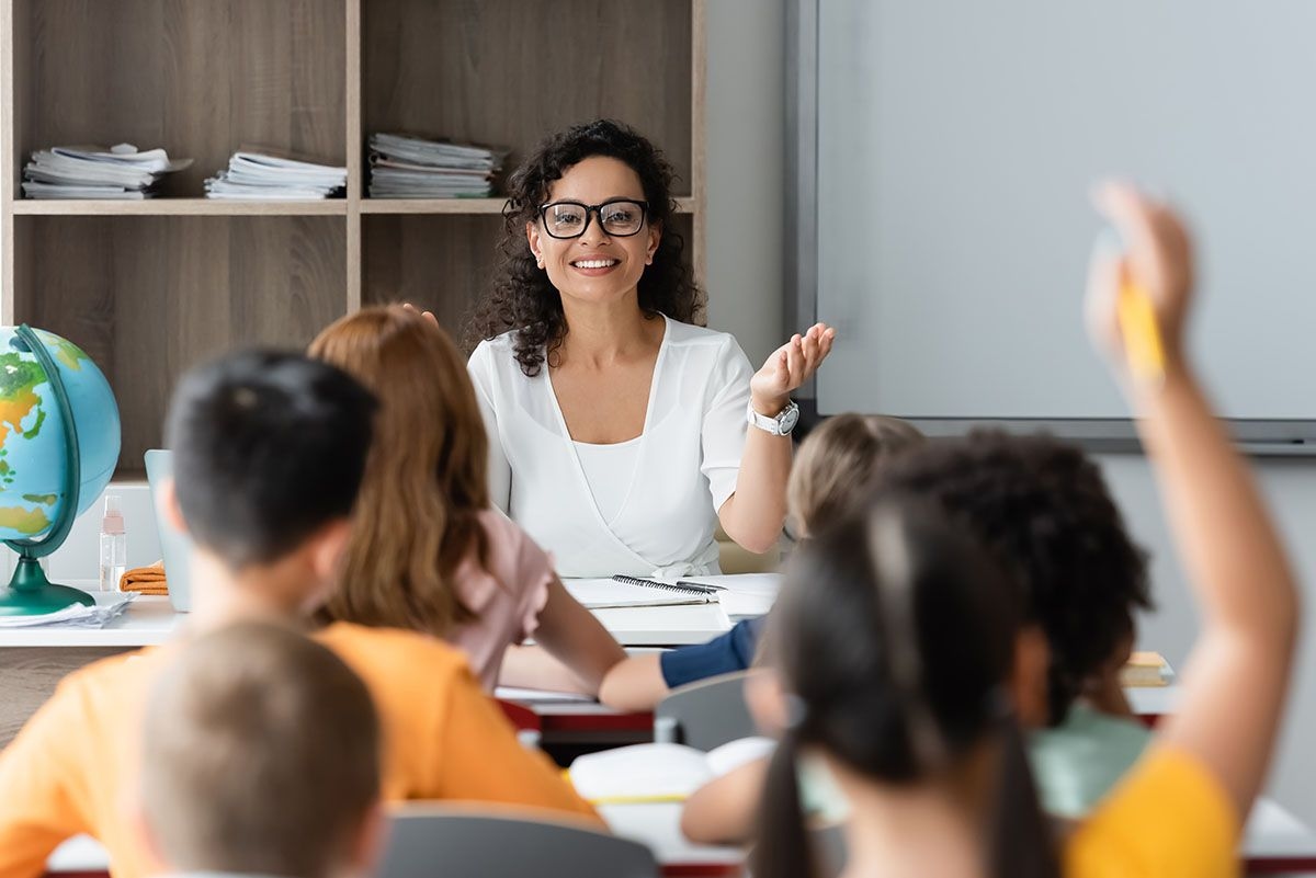Eine glückliche Lehrerin sitzt vor einer Schulklasse. Sie nimmt ein Kind dran, das sich gerade meldet.